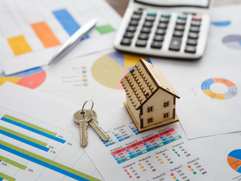 Miniatura de casa sobre gráficos financeiros, chaves e calculadora ao fundo. Quanto preciso dar de entrada para financiar um apartamento?