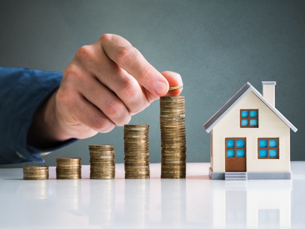 Pilhas de moedas crescendo ao lado de uma casa em miniatura, ilustrando investimentos imobiliários. Imóvel é um bom investimento? Entenda agora!