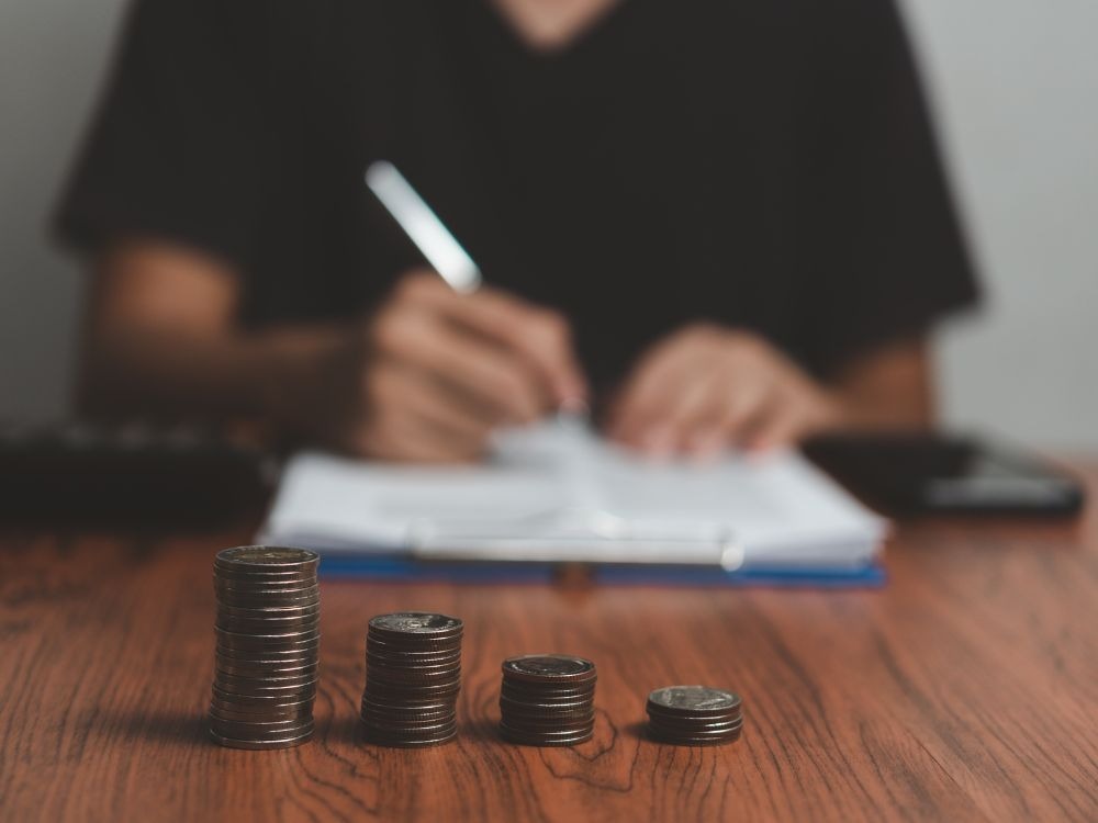 Moedas empilhadas em destaque enquanto uma pessoa faz cálculos ao fundo. Imóvel é um bom investimento? Entenda agora!