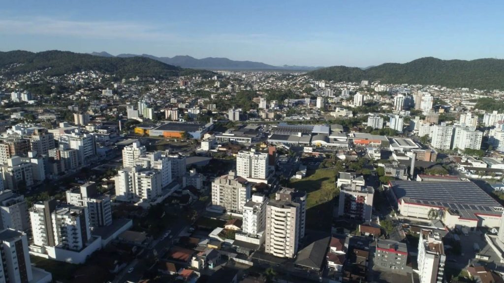 Vista aérea com prédios residenciais, áreas comerciais e infraestrutura urbana do Bairro América em Joinville ao fundo.