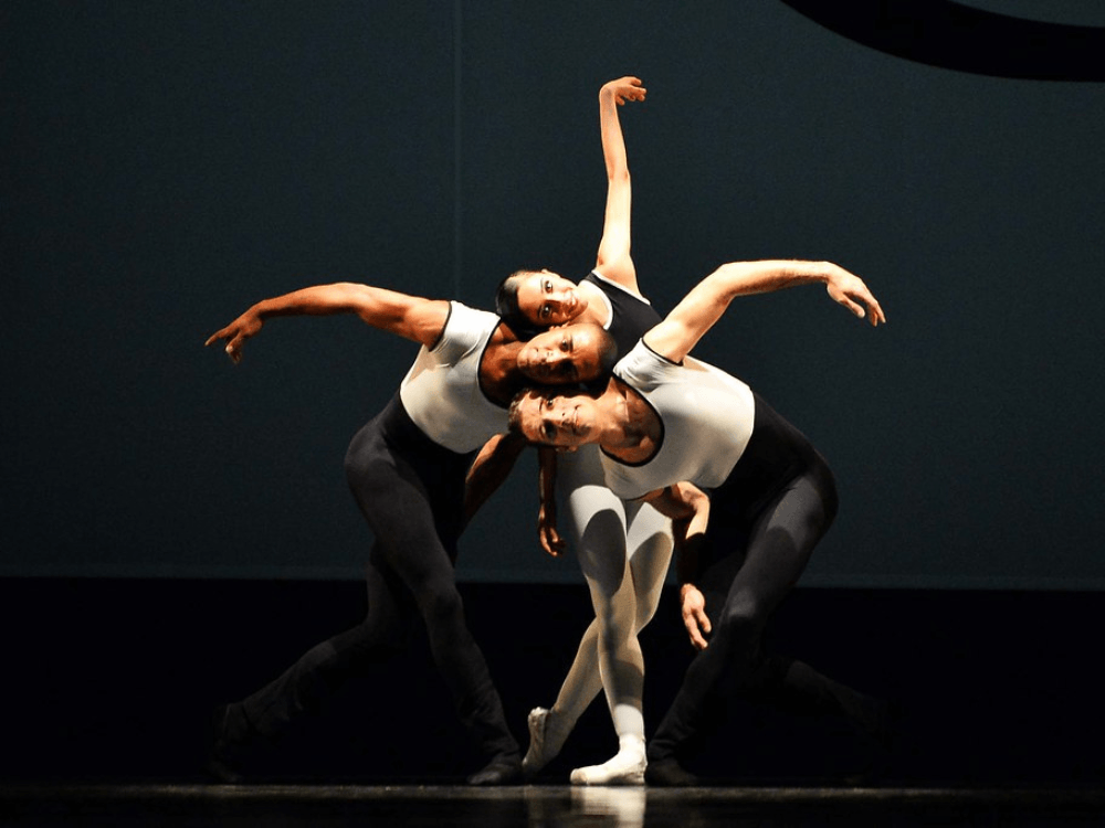Três bailarinos executam uma composição de dança contemporânea com movimentos integrados durante espetáculo em palco.