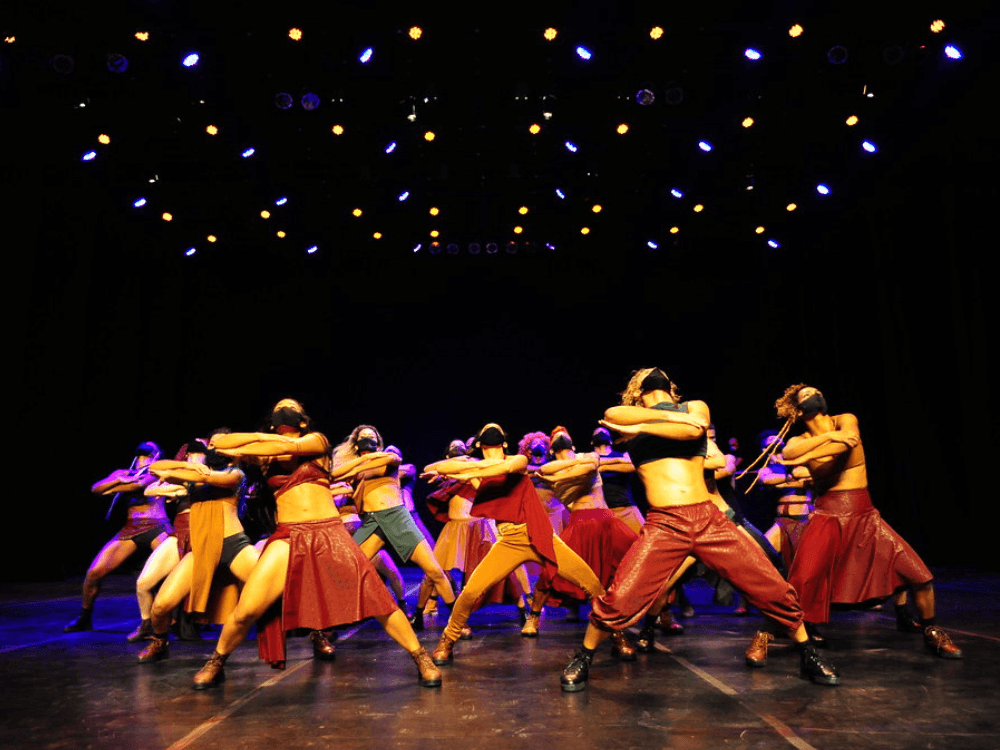 Companhia de dança realiza apresentação coletiva com figurinos coloridos e iluminação cênica em espetáculo do festival.