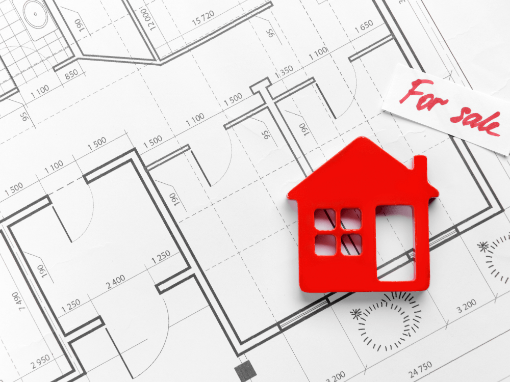 Planta arquitetônica com pequena casa vermelha e placa de venda representa a escolha de um imóvel para comprar. Tudo para mostrar se é melhor comprar imóvel na planta ou pronto.