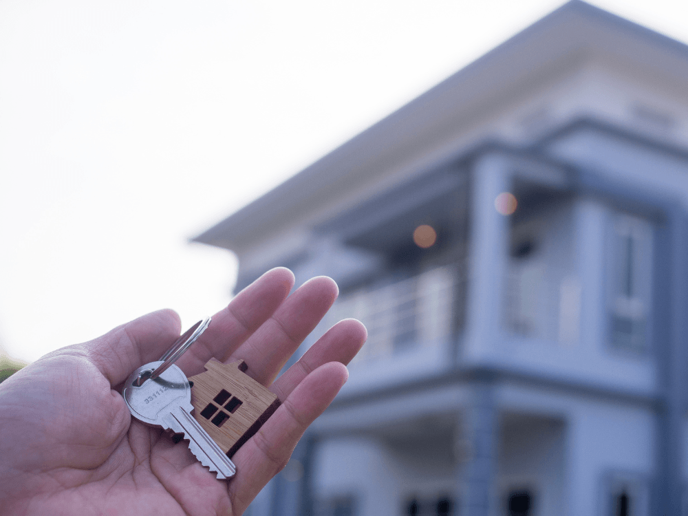 Mão segura chaves e uma pequena casa de madeira diante de um imóvel, simbolizando a compra da casa própria em Joinville.
