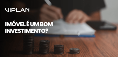 Pilhas de moedas sobre a mesa com pessoa escrevendo ao fundo, destacando análise financeira. Imóvel é um bom investimento? Entenda agora!